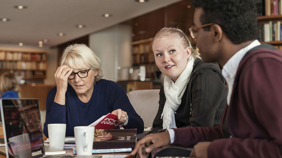 Tre studenter i samtal under ett grupparbete på universitetsbibliotek.