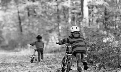 Två barn i cykelhjälm som leder sina cyklar längs vägkanten intill en skog, med ryggarna mot kameran. Marken är täckt av höstlöv.