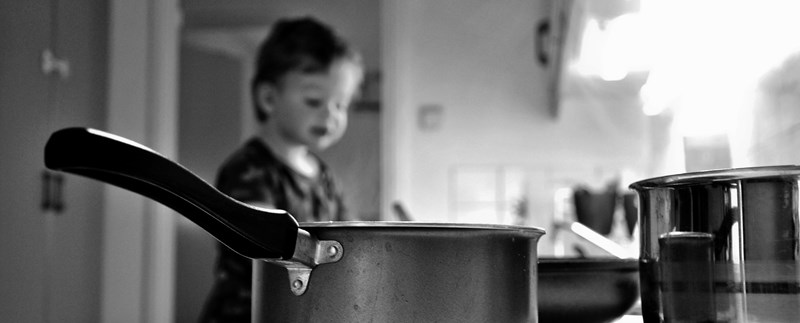 Litet barn i ett kök med kastruller på spisen i förgrunden.