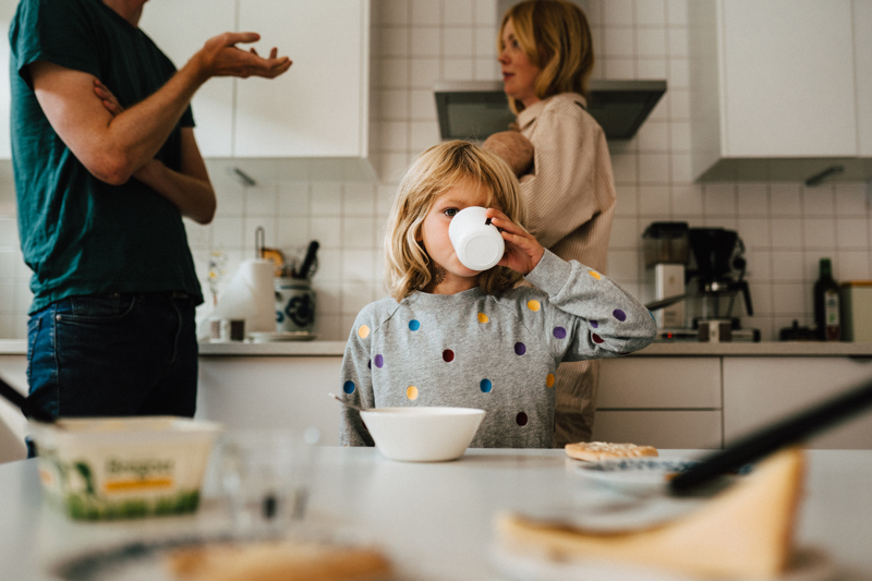 En flicka sitter vid ett frukostbord och dricker ur en mugg framför två vuxna som diskuterar och gestikulerar i köket. Foto: Stina Gränfors