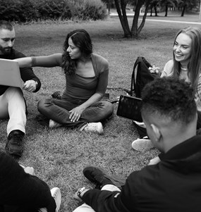 Sex personer sitter i ring på gräsmatta och diskuterar något som visas på laptop.