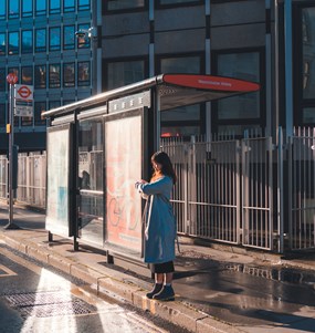 En kvinna som väntar på en buss vid en busshållsplats. Solen skiner men det har nyss regnat.