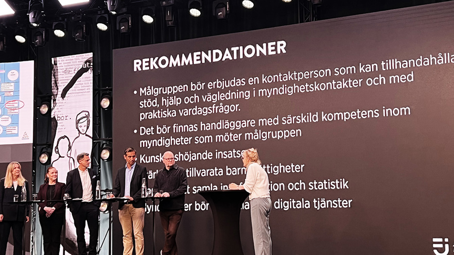 Sex personer på en sen framför talarbord. I bakgrunden visas en powerpoint med rubriken "Rekommendationer" och en punktlista med text som inte är helt läsbar.