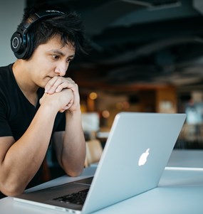 En man med hörlurar som ser koncentrerat på sin datorskärm. Han sitter i en modern miljö, kontor eller ett café. 