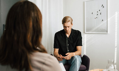 Ung man i samtal med kvinna som bara syns bakifrån. Mannen ser ner på sina händer. Miljön liknar rum för samtalsterapi..