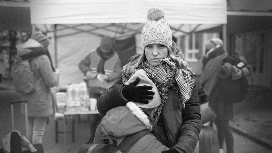 Ukrainsk kvinna står vid ett tält där vatten delas ut. Hon tittar allvarligt in i kameran medan hennes barn ledset trycker pannan mot kvinnan. Kvinnan omfamnar barnet.