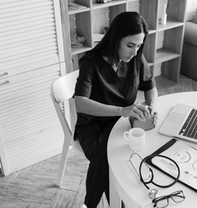 En kvinna i vårdpersonal-klädsel sitter vid ett bord framför en dator och tittar ner i sin plånbok. På bordet ligger ett stetoskop, ett par glasögon och papper med olika diagram