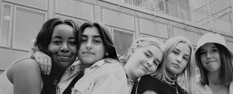 Fem unga tjejer som håller om varandra framför en betongbyggnad