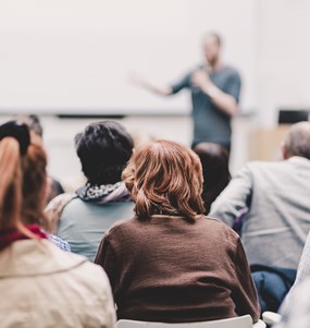 Vuxna studenter sett bakifrån som lyssnar till en manlig lärare i oskärpa vid en whiteboard