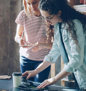En kvinna står vid ett arbetsbord med en kamera. En annan kvinna står bakom henne och för anteckningar.