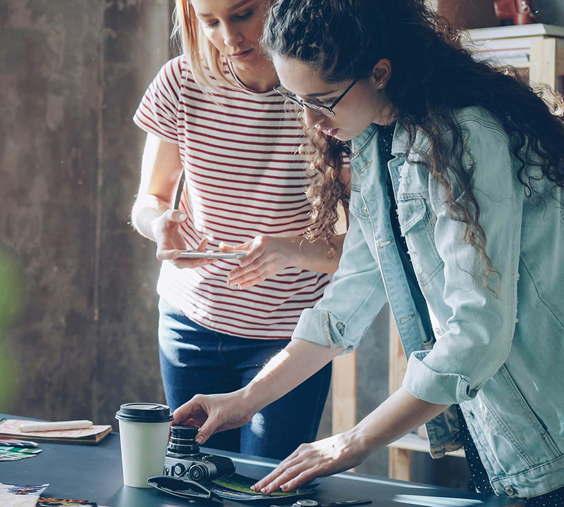 En kvinna står vid ett arbetsbord med en kamera. En annan kvinna står bakom henne och för anteckningar.