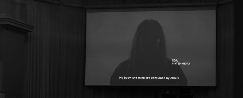 Ett videoklipp spelas upp i en konferenssal. Bilden visar en kvinna i skuggan vars ansikte inte syns. Hon säger "My body isn't mine. It's consumed by others". IDA #INTEDINHORA