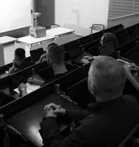 En sluttande föreläsningssal i universitetsmiljö där studenter sitter i åhörarstolar och en kvinna står längst fram och talar. 