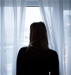 Kvinna står och tittar ut genom ett fönster bortvänd från kameran