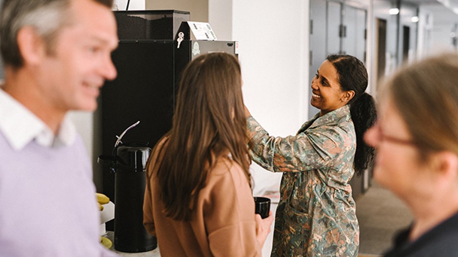 Fyra personer samlade runt kaffeautomaten i ett personalrum. De är i samtal två och två och håller kaffekoppar i händerna. 