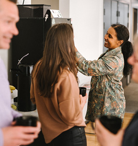 Fyra personer samlade runt kaffeautomaten i ett personalrum. De är i samtal två och två och håller kaffekoppar i händerna.