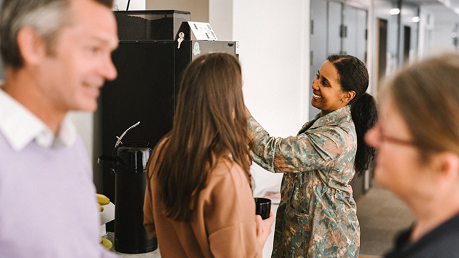 Fyra personer samlade runt kaffeautomaten i ett personalrum. De är i samtal två och två och håller kaffekoppar i händerna.