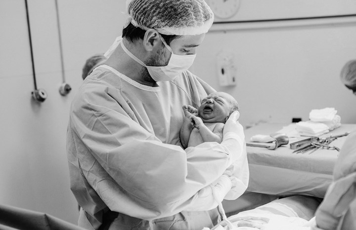 En man i sjukhuskläder och munskydd håller en helt nyfödd bebis i famnen. Navelsträngen är fortfarande kvar. I bakgrunden syns typisk sjukhusmiljö. 