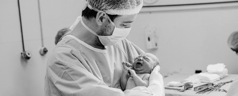 En man i sjukhuskläder och munskydd håller en helt nyfödd bebis i famnen. Navelsträngen är fortfarande kvar. I bakgrunden syns typisk sjukhusmiljö. 