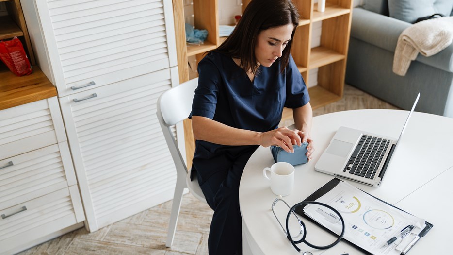 Kvinna i sjukvårdskläder sitter vid dator och öppnar sin plånbok.