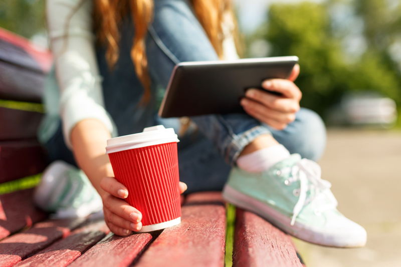 Ung kvinna sitter på en parkbänk med en kaffekopp och tittar på en surfplatta. Foto: Mostphotos