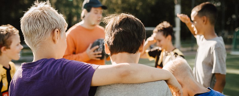 En manlig tränare pratar med unga pojkar i ett fotbollslag. Två barn är i förgrunden där den ena lägger armen om den andra.