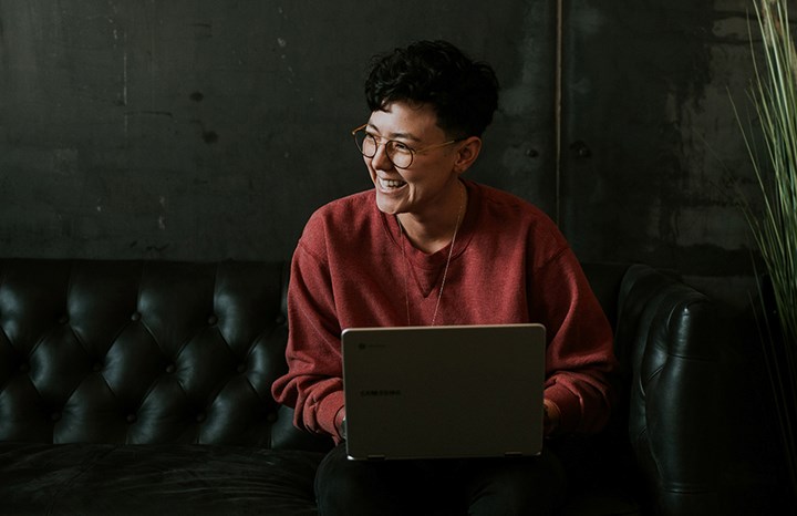 En person i en mörk soffa som leende ser upp från sin laptop och tittar åt sidan. Miljön är mörk. Personen är klädd i röd tröja och glasögon och intill soffan finns en stor grön växt. 